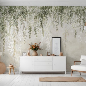 May include: A room with a white floor and a white cabinet. The wall features a mural of green and white hanging leaves. A white armchair with wooden arms and a small side table with a lamp are also present. A vase of orange flowers sits on the cabinet.