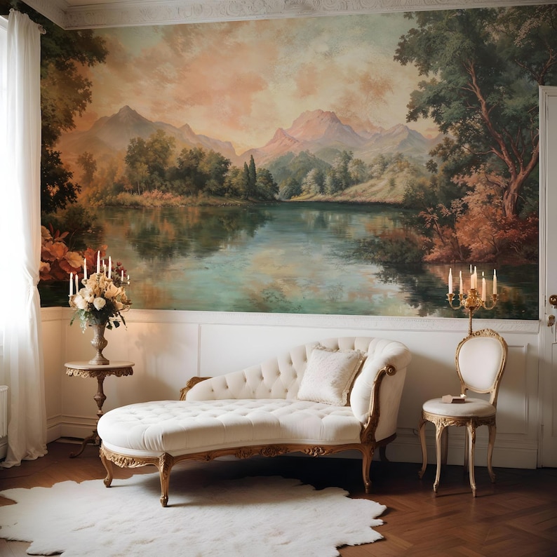 May include: A white chaise lounge with gold accents sits in a room with a large mural depicting a lake and mountains. The mural is painted in shades of green, blue, and brown. A white chair with gold accents and a small table with a book on it are also in the room.