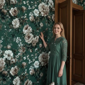 May include: A woman in a green dress stands next to a wall covered in floral wallpaper. The wallpaper features large, cream-colored peony blooms and smaller white flowers against a dark green background. A matching floral pillow is visible on a nearby couch.