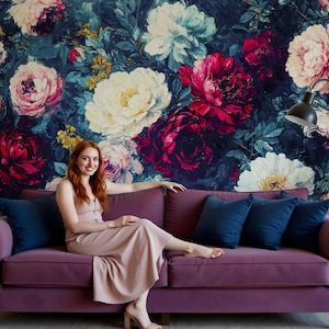 May include: A woman sits on a purple sofa with navy blue cushions. The sofa is in front of a wall covered in a floral wallpaper with large blooms in shades of pink, white, and red.