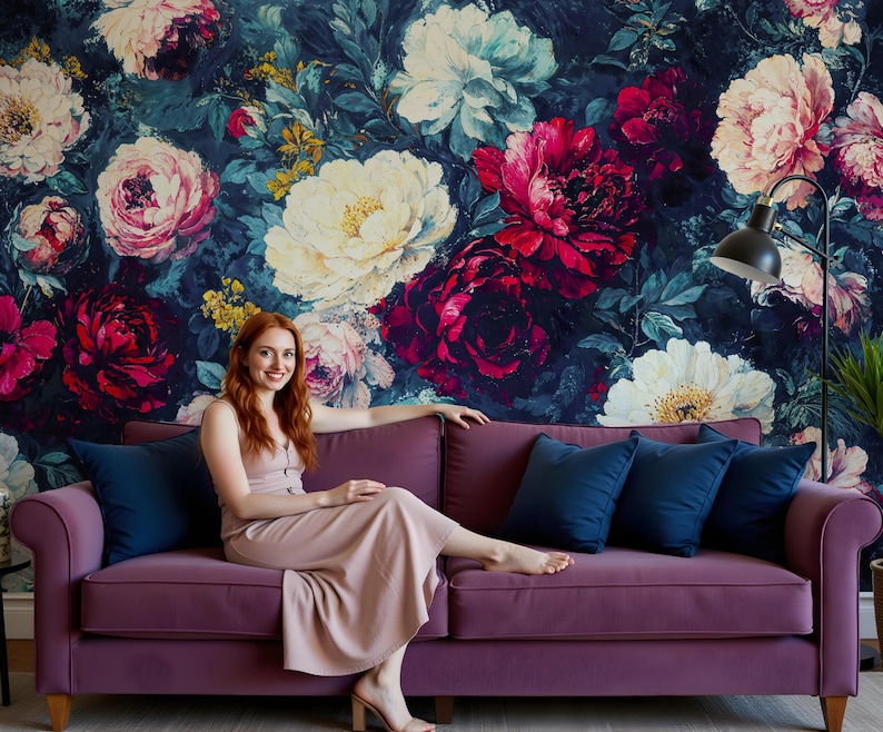 May include: A woman sits on a purple sofa with navy blue cushions. The sofa is in front of a wall covered in a floral wallpaper with large blooms in shades of pink, white, and red.