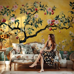 May include: A woman sits on a floral patterned sofa in front of a yellow wall with a large painted floral tree. The tree has pink flowers and red apples.