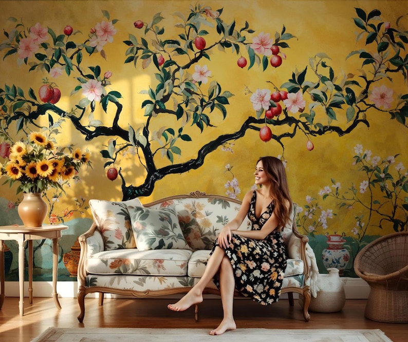 May include: A woman sits on a floral patterned sofa in front of a yellow wall with a large painted floral tree. The tree has pink flowers and red apples.