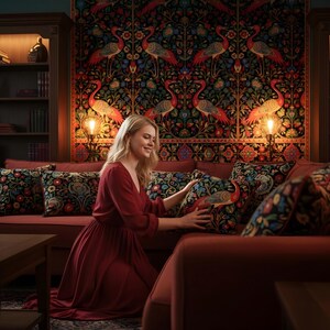 May include: A living room scene with a red sofa and decorative pillows featuring floral and bird designs. A woman in a red dress is arranging the pillows. The room has a dark wood bookcase and a patterned tapestry on the wall.
