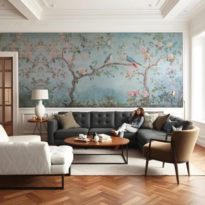May include: A living room with a sectional sofa in dark grey fabric, a white tufted armchair, a round wooden coffee table, and a floral wallpaper mural featuring a tree with birds.