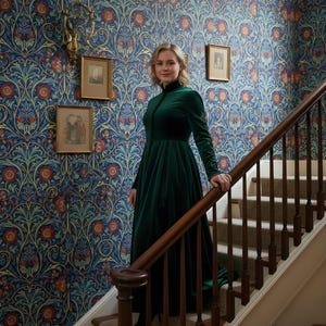 May include: A woman in a long, dark green velvet dress stands on a staircase with a wooden banister. The dress has long sleeves and a high collar. The wallpaper behind her features a blue floral pattern with red and orange accents. Small framed pictures hang on the wall.