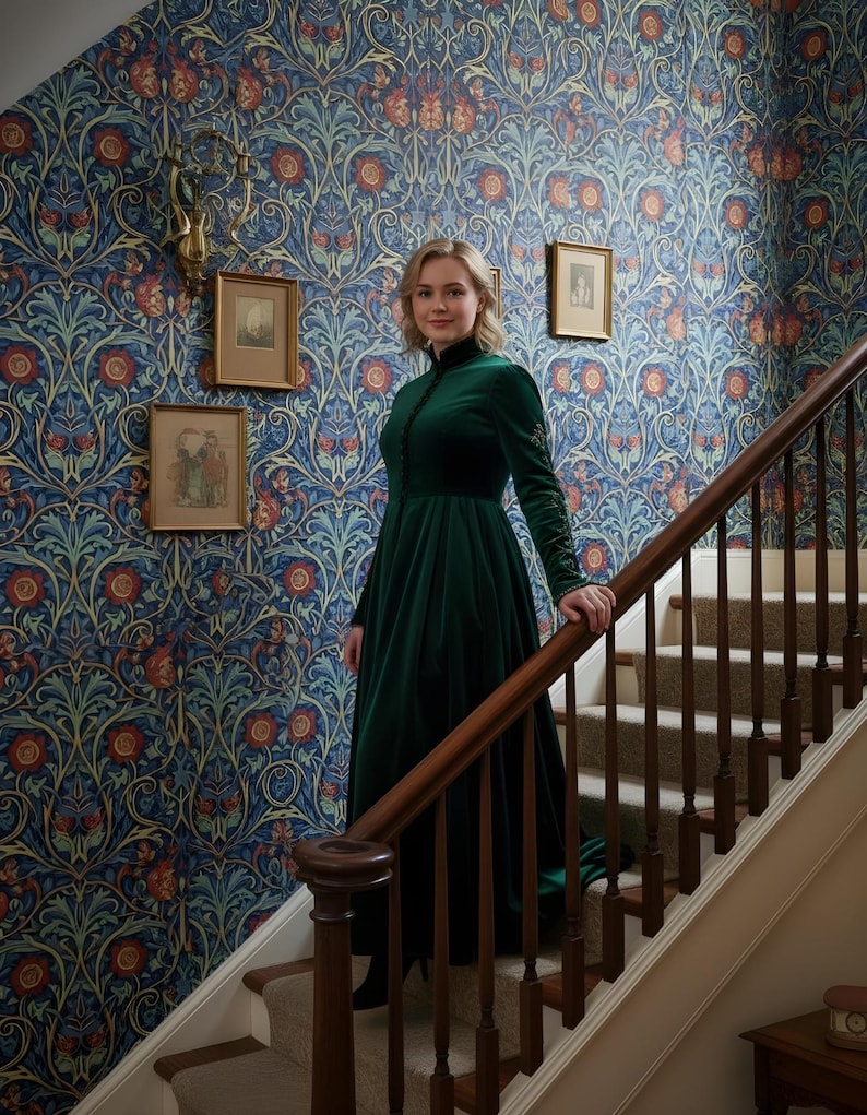 May include: A woman in a long, dark green velvet dress stands on a staircase with a wooden banister. The dress has long sleeves and a high collar. The wallpaper behind her features a blue floral pattern with red and orange accents. Small framed pictures hang on the wall.