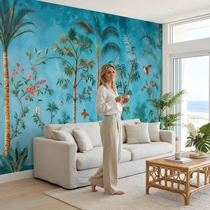 May include: A living room with a vibrant blue mural featuring palm trees, flowers, and butterflies. A beige sofa with pillows, a rattan coffee table, and a woman in a white shirt and beige pants are in the room.