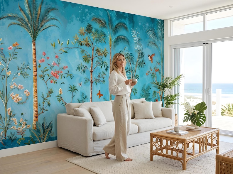 May include: A living room with a vibrant blue mural featuring palm trees, flowers, and butterflies. A beige sofa with pillows, a rattan coffee table, and a woman in a white shirt and beige pants are in the room.
