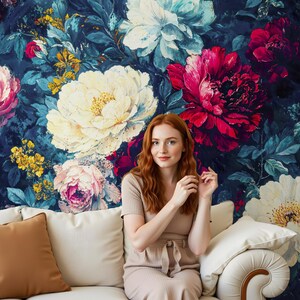 May include: A woman sits on a white sofa in front of a floral wallpaper. The wallpaper features large, colorful flowers in shades of white, pink, and red against a dark blue background. The sofa has a brown pillow and a white pillow.