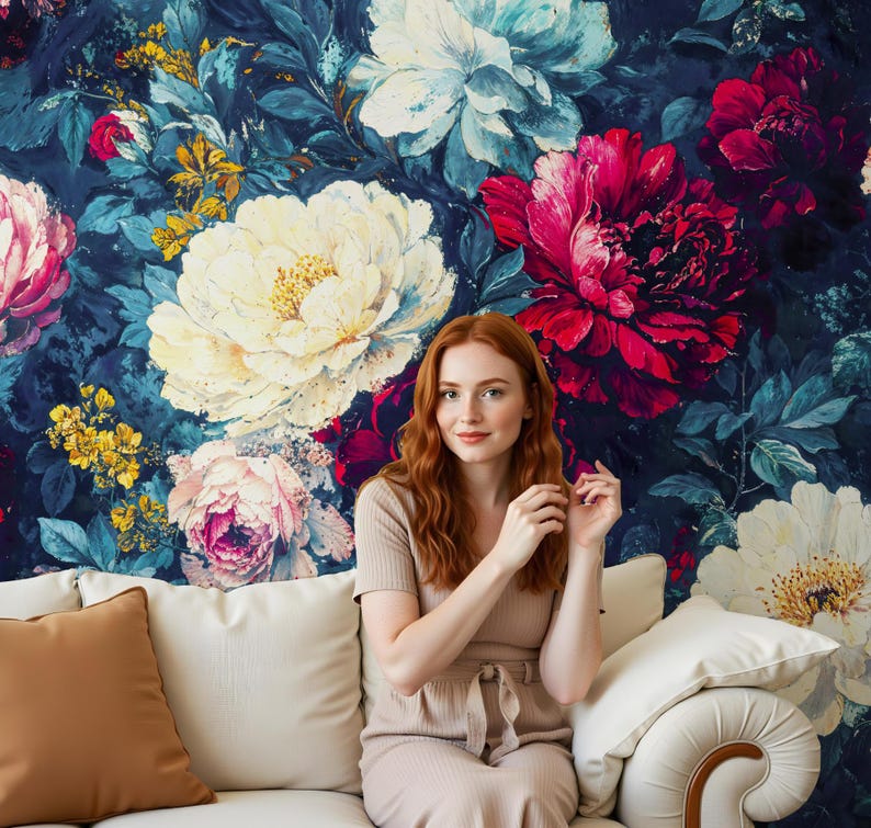 May include: A woman sits on a white sofa in front of a floral wallpaper. The wallpaper features large, colorful flowers in shades of white, pink, and red against a dark blue background. The sofa has a brown pillow and a white pillow.