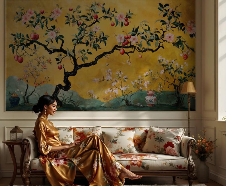 May include: A woman in a gold silk robe sits on a floral patterned sofa in front of a wall mural depicting a flowering tree with red fruit. The mural is painted in shades of yellow, green, and white.