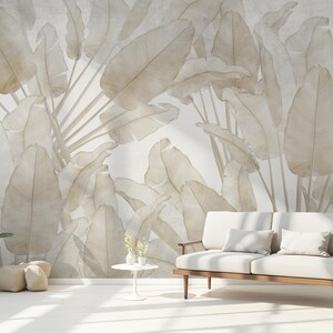 May include: A neutral-toned interior scene featuring a large-scale mural of stylized banana leaves in shades of beige and cream. A white sofa with wooden arms and legs is in front of the mural. A potted plant and small side table complete the room.