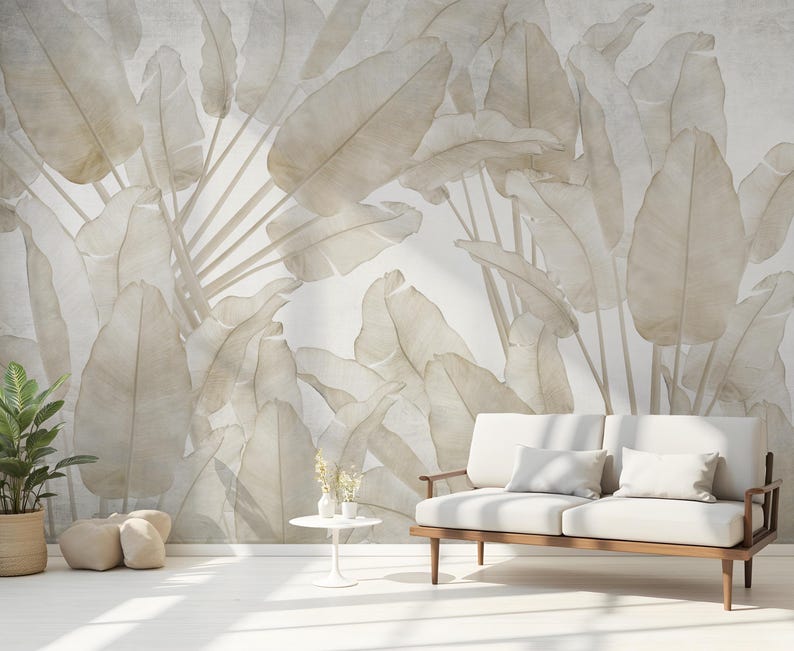 May include: A neutral-toned interior scene featuring a large-scale mural of stylized banana leaves in shades of beige and cream. A white sofa with wooden arms and legs is in front of the mural. A potted plant and small side table complete the room.