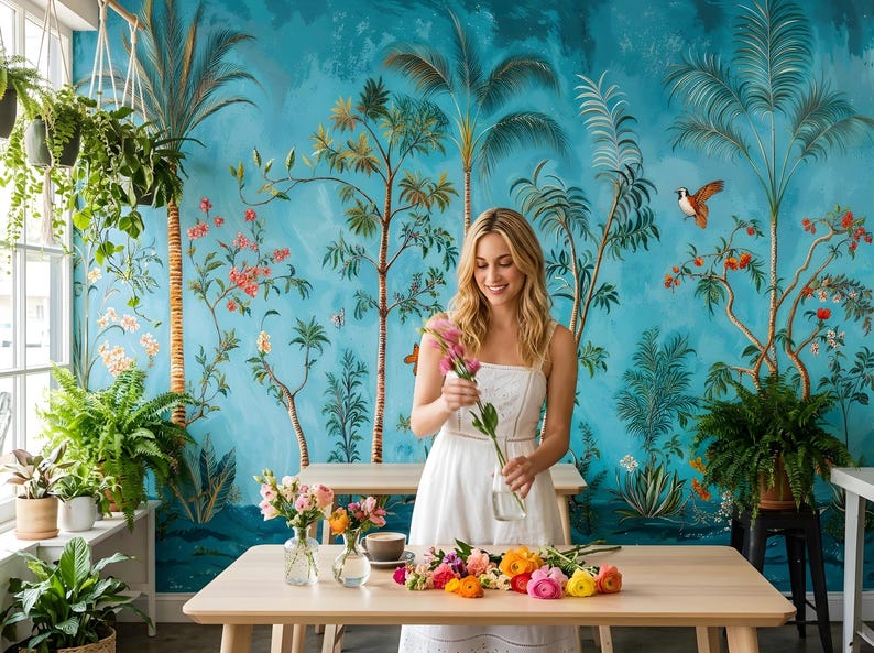 May include: A woman in a white dress arranging flowers in a vase. The scene is set in a room with a tropical mural on the wall. A table is covered with colorful flowers. Plants and greenery are visible.