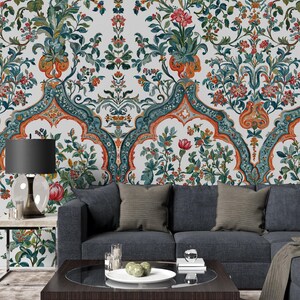 Op de afbeelding: Een gedetailleerd bloemenbehangontwerp met een herhalend patroon van bloemen en bladeren in tinten rood, groen, oranje en blauwgroen. Het behang is ge&iuml;nstalleerd op een muur achter een donkergrijze bank met decoratieve kussens.