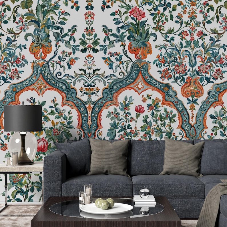 Op de afbeelding: Een gedetailleerd bloemenbehangontwerp met een herhalend patroon van bloemen en bladeren in tinten rood, groen, oranje en blauwgroen. Het behang is ge&iuml;nstalleerd op een muur achter een donkergrijze bank met decoratieve kussens.