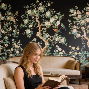 May include: A woman reading a book on a beige sofa in front of a black floral wallpaper. The wallpaper features flowering trees with birds in shades of white, pink, and green. The room has dark wood furniture and a traditional aesthetic.