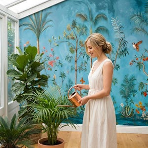 May include: A woman in a white dress waters plants in a sunroom. The room features a blue mural with palm trees and birds. A copper watering can is used on a potted plant. Other plants are in the foreground, and a wicker chair is in the background.
