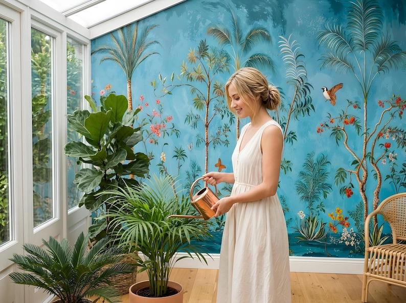 May include: A woman in a white dress waters plants in a sunroom. The room features a blue mural with palm trees and birds. A copper watering can is used on a potted plant. Other plants are in the foreground, and a wicker chair is in the background.