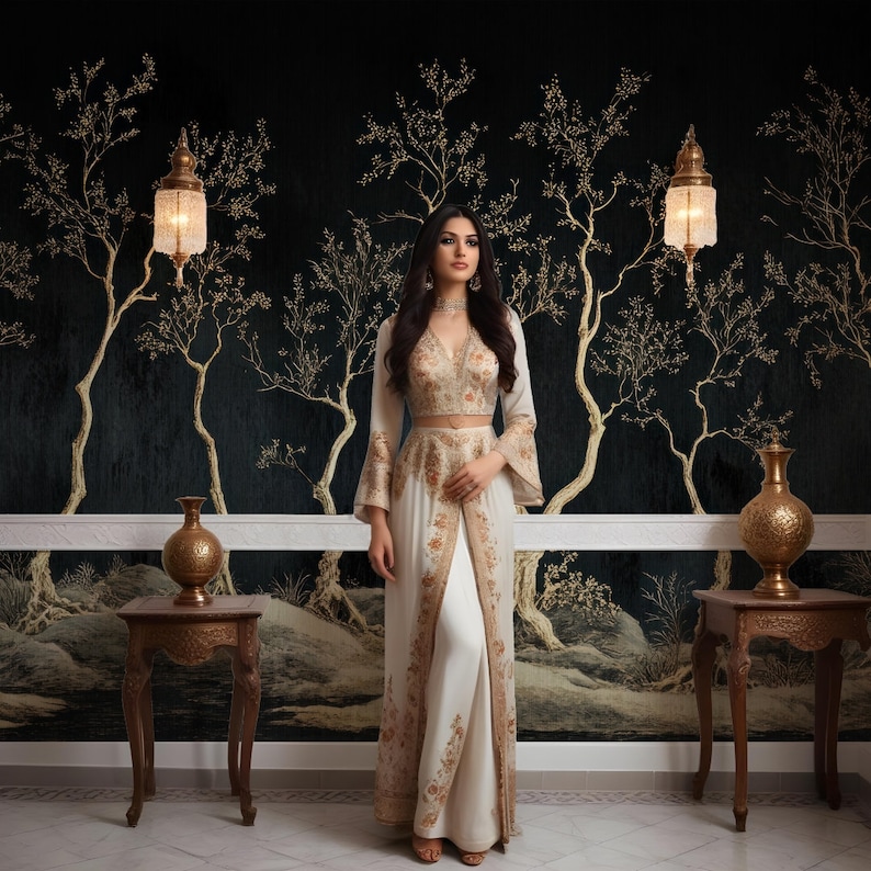 May include: A woman wearing a white and gold embroidered outfit with a long skirt and a matching top. She is standing in front of a black and gold wallpaper with a tree design.