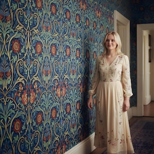 May include: A woman in a cream-colored dress with floral embroidery stands in a hallway. The dress has long sleeves and a V-neck. The wall behind her is covered in a blue floral wallpaper with red and green accents. A patterned rug is on the floor.