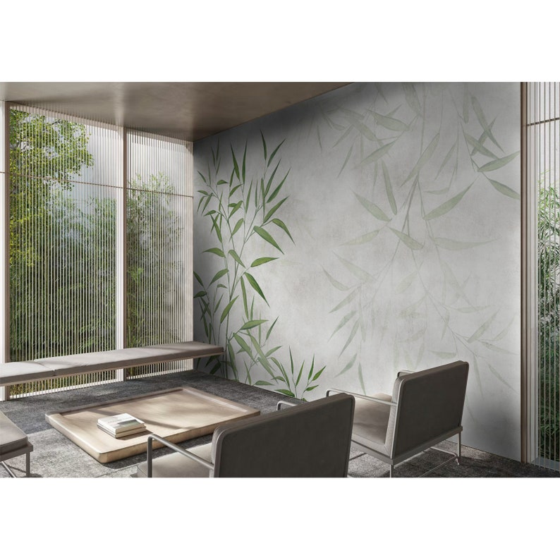 Op de afbeelding: Een woonkamer met een lichtgrijze muur met een groen bamboe blad behangontwerp. Twee grijze stoelen staan voor de muur, en een salontafel met een boek erop staat in het midden van de kamer.