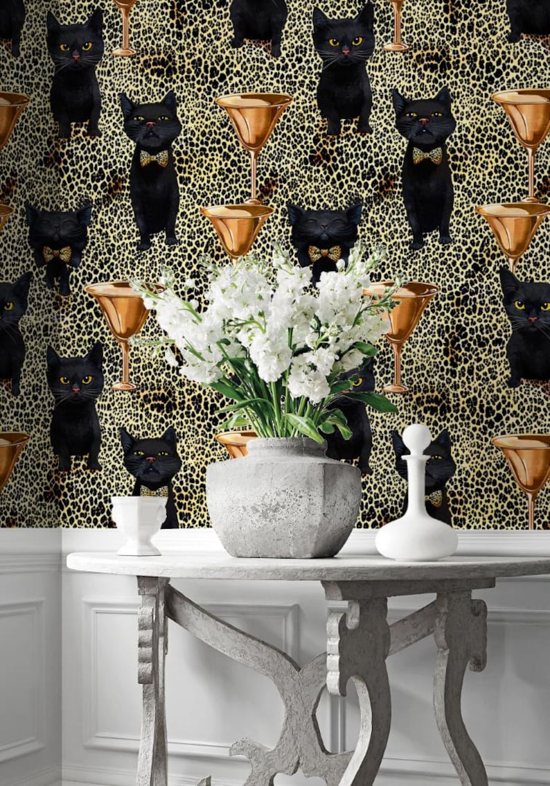May include: A gray console table with a black and gold wallpaper featuring a repeating pattern of black cats wearing bow ties and martini glasses. White flowers are in a gray ceramic vase on the table.