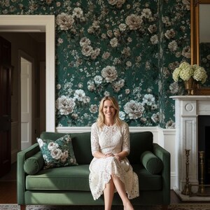 May include: A woman sits on a green velvet sofa in a room with floral wallpaper. She wears a white lace dress and nude heels. A matching floral pillow rests on the sofa. The room features a fireplace and a gold-framed mirror.