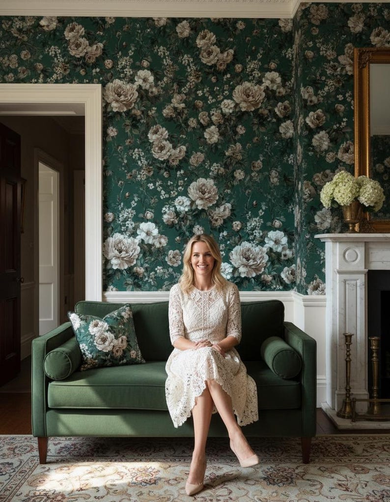 May include: A woman sits on a green velvet sofa in a room with floral wallpaper. She wears a white lace dress and nude heels. A matching floral pillow rests on the sofa. The room features a fireplace and a gold-framed mirror.
