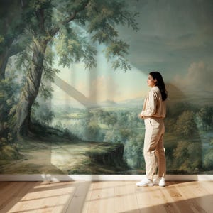 May include: A woman in a beige outfit stands in front of a large landscape mural. The mural depicts a lush green forest with a river winding through it. The woman is looking off to the side, and the light from a window is casting a shadow on the wall behind her.