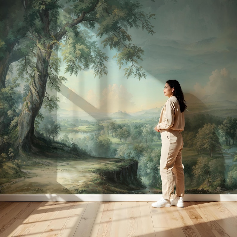 May include: A woman in a beige outfit stands in front of a large landscape mural. The mural depicts a lush green forest with a river winding through it. The woman is looking off to the side, and the light from a window is casting a shadow on the wall behind her.