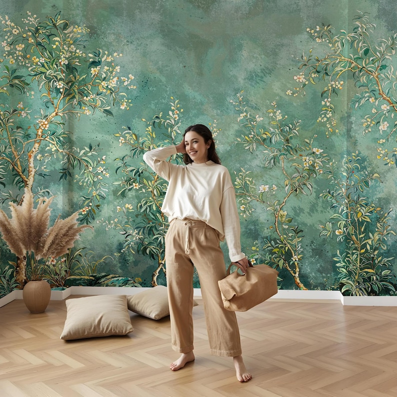 May include: A woman stands in front of a green floral wallpaper with white flowers. She is wearing a white long-sleeved shirt and brown wide-leg trousers. She is holding a brown tote bag.