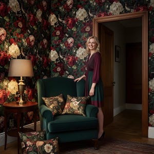 May include: A room with floral wallpaper in shades of red, cream, and green. A dark green armchair with floral pillows sits in front of the wall. A woman stands in the doorway. A round side table with a lamp is also visible.