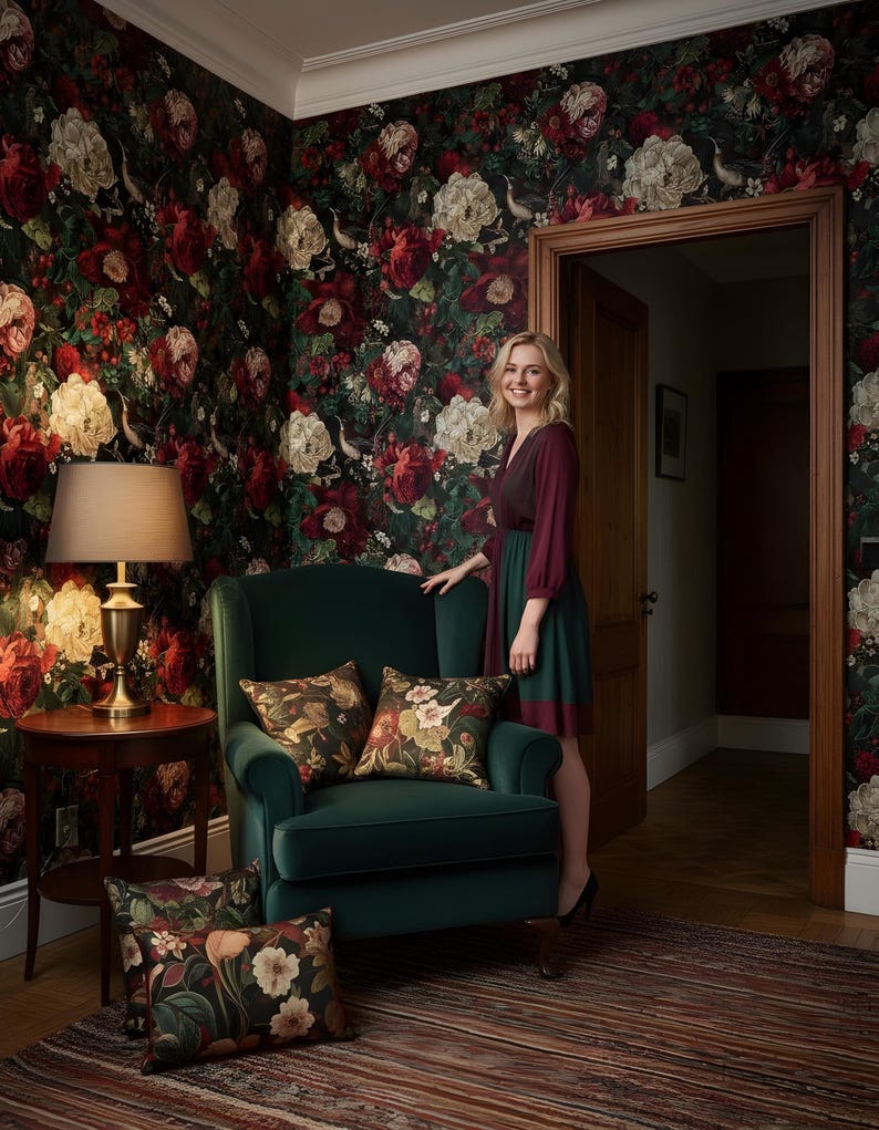 May include: A room with floral wallpaper in shades of red, cream, and green. A dark green armchair with floral pillows sits in front of the wall. A woman stands in the doorway. A round side table with a lamp is also visible.
