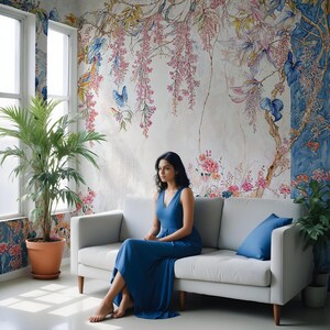 May include: A woman in a blue dress sits on a light gray sofa in front of a floral mural. The mural features pink, blue, and green flowers and vines. A potted plant sits to the left of the sofa, and a blue pillow rests on the sofa.