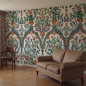 Op de afbeelding: Een kamer met bloemenbehang in cr&egrave;me, rood, groen en oranje. Een bruine leren bank en een houten stoel staan in de kamer. Het behang heeft een herhalend bloemenpatroon.