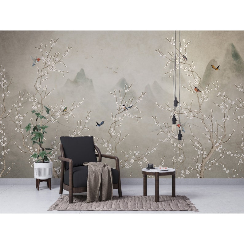 Op de afbeelding: Een grijze en witte bloemige muurschildering met een berglandschap en vogels. Een zwarte fauteuil met een deken zit voor de muurschildering, en een kleine ronde tafel met een wit blad staat naast de stoel.