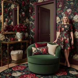 May include: A room with floral wallpaper and a matching rug. A green swivel chair has decorative pillows. A woman in a floral dress stands near the chair. A gold-framed mirror and a side table with a vase of flowers are also visible.