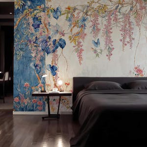 May include: A bedroom with a floral mural featuring blue, pink, and green botanical elements. The mural covers the wall behind a bed with dark gray bedding. A small black side table with a lamp and decor sits next to the bed.
