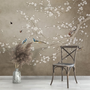 May include: A brown wooden chair with a cross-back design sits in front of a wall with a floral wallpaper featuring a delicate branch of white blossoms and blue birds.