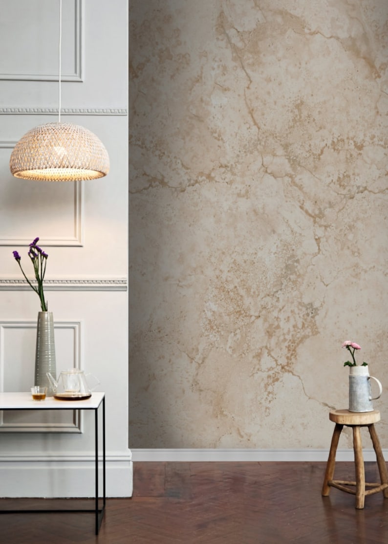 Op de afbeelding: Een beige, gestructureerd behang met een lichtbruin en wit marmerpatroon. Het behang bevindt zich in een kamer met een wit plafond en witte muren. Op de voorgrond staat een witte tafel met een zwart metalen frame. Een hanglamp van wilgentakken hangt aan het plafond.