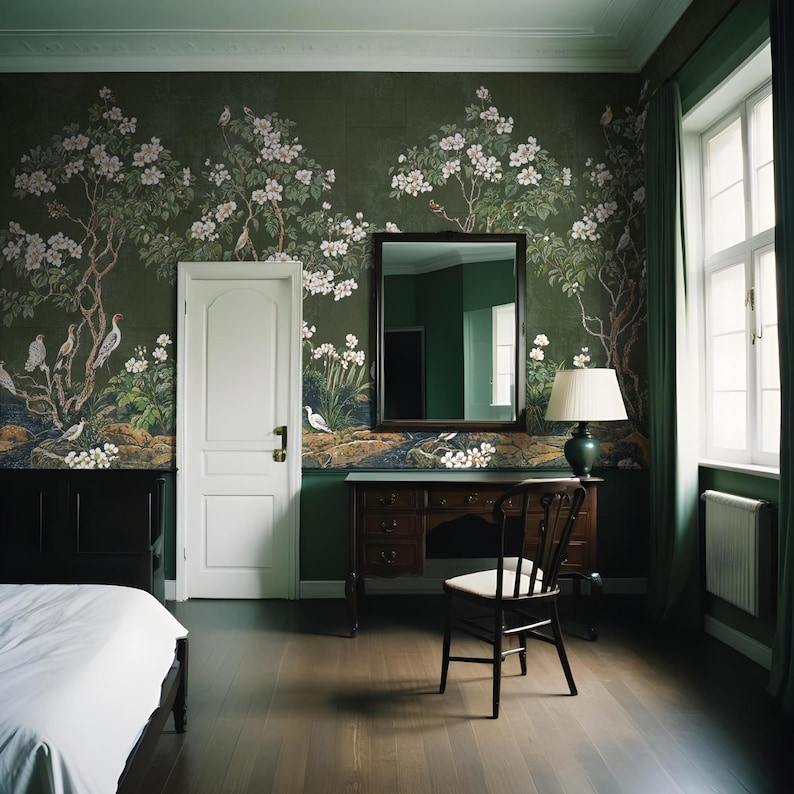 May include: A bedroom interior with a green wall featuring a floral wallpaper design. A white bed with a white duvet is in the foreground. A wooden desk with a chair is in the middle of the room. A mirror hangs on the wall above the desk.