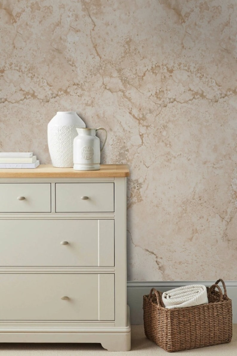 Op de afbeelding: Een witte commode met vier lades en een houten blad. Een witte vaas en een witte kan met een bloemenpatroon staan op de commode. Een bruine rieten mand met witte handdoeken staat op de vloer voor de commode. De muur achter de commode is een beige marmerbehang.