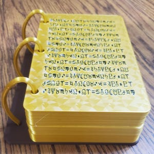 May include: A gold-colored, multi-page book with a ring binding. The pages are filled with black text and symbols. The book is on a wooden surface.