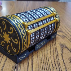 May include: A black and gold Cryptex cipher cylinder with rotating lettered rings. The ends feature decorative patterns, and the base is black. The word "Cryptex" is printed on the base.