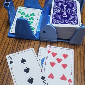 May include: A blue plastic card holder with two compartments, one holding a deck of cards and the other holding a card with the number 10 and green clover symbols. Three playing cards are in front.