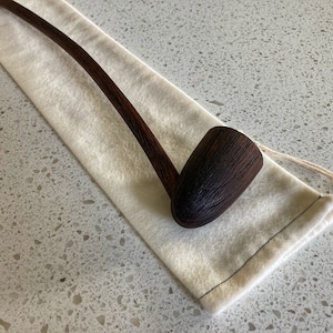 Puede incluir: Una pipa larga de madera marrón oscura con una boquilla curva y una cazoleta redonda. La pipa está apoyada en una bolsa de tela blanca.