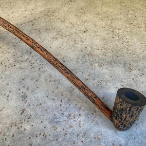 May include: A wooden tobacco pipe with a long, slender stem and a rounded bowl. The pipe is made of dark brown wood with a textured grain pattern. The bowl has a dark, almost black, finish.