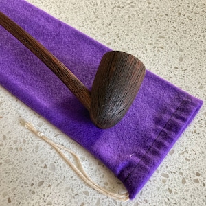 Puede incluir: Una pipa de madera marrón oscuro con una larga boquilla que descansa sobre una bolsa de terciopelo morado con un cordón de ajuste blanco.